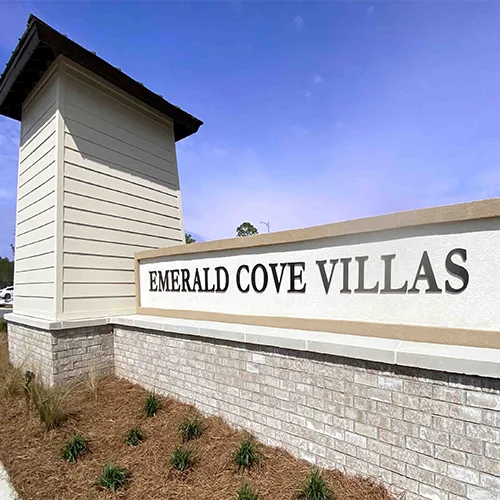 Entrance monument sign for EMERALD COVE VILLAS with beige siding tower and brick base under a blue sky
